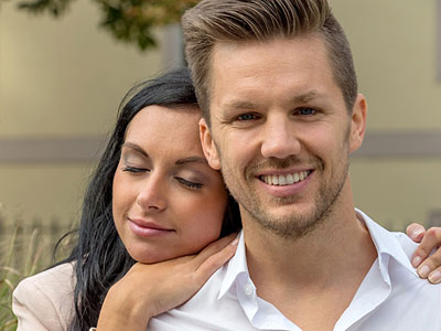 Ein lächelndes Paar steht im Freien, die Frau umarmt den Mann liebevoll.